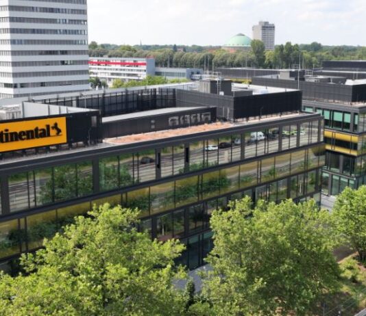 Das Bild zeigt ein modernes Bürogebäude mit dem „Continental“-Logo. Umgeben von Glasgebäuden, Grünflächen und Bäumen, ist im Hintergrund ein hoher weißer Bau und ein entferntes Wahrzeichen mit grüner Kuppel sichtbar. Eine Straße verläuft entlang des Komplexes, mit einigen Fahrzeugen, und der Himmel ist teils bewölkt.