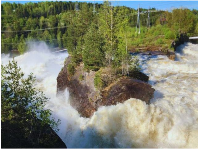 Das Bild zeigt einen kraftvollen Wasserfall, der stark über eine große, felsige Formation mit Bäumen stürzt. Umgeben von dichten grünen Wäldern und einem klaren Himmel, sind im Hintergrund auch Stromleitungen und Versorgungsinfrastruktur sichtbar, was eine Mischung aus Natur und menschlichem Einfluss zeigt.