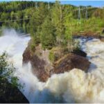 Das Bild zeigt einen kraftvollen Wasserfall, der stark über eine große, felsige Formation mit Bäumen stürzt. Umgeben von dichten grünen Wäldern und einem klaren Himmel, sind im Hintergrund auch Stromleitungen und Versorgungsinfrastruktur sichtbar, was eine Mischung aus Natur und menschlichem Einfluss zeigt.