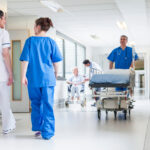 Symbolbild Krankenhaus (Male nurse pushing stretcher gurney bed in hospital corridor with doctors & senior female patient.) Copyright: spotmatikphoto - stock.adobe.com