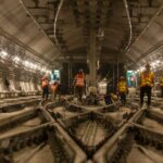 Das Bild zeigt eine Szene in einem großen unterirdischen Tunnel. Mehrere Bauarbeiter, die orange und gelbe reflektierende Sicherheitswesten und Helme tragen, sind im Tunnel verteilt und mit verschiedenen Aufgaben beschäftigt. Der Tunnel wird von zahlreichen Lichtern entlang der Wände beleuchtet, und in der Mitte verlaufen mehrere Bahngleise. An den Seiten sind Kabel und andere Baumaterialien zu sehen, die zu einem aktiv bearbeiteten Infrastrukturprojekt gehören.