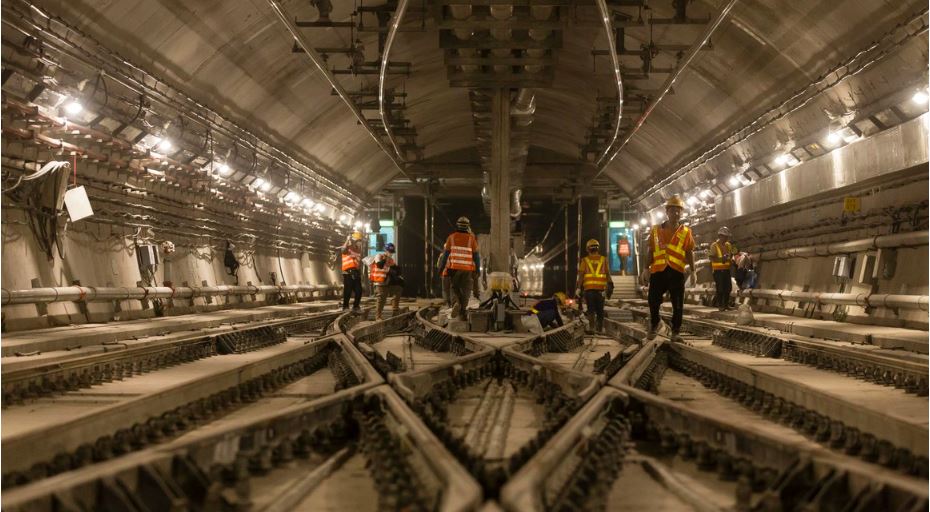 Das Bild zeigt eine Szene in einem großen unterirdischen Tunnel. Mehrere Bauarbeiter, die orange und gelbe reflektierende Sicherheitswesten und Helme tragen, sind im Tunnel verteilt und mit verschiedenen Aufgaben beschäftigt. Der Tunnel wird von zahlreichen Lichtern entlang der Wände beleuchtet, und in der Mitte verlaufen mehrere Bahngleise. An den Seiten sind Kabel und andere Baumaterialien zu sehen, die zu einem aktiv bearbeiteten Infrastrukturprojekt gehören.